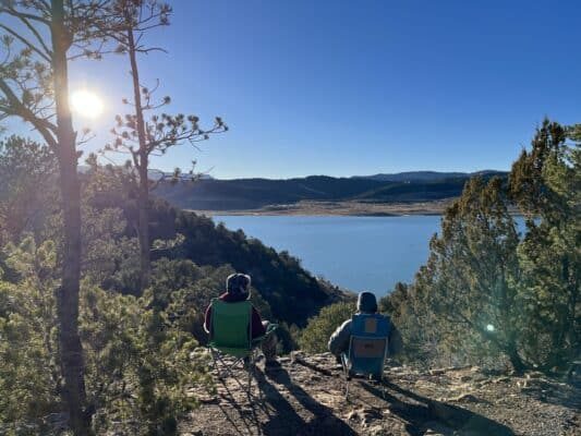 Two campers at Trinidad Lake State Park Campground sit in camp chairs and sip coffee. They sit above the lake and admire the view of Fisher's Peak in the distance.